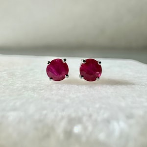 Puede incluir: Un par de pendientes de botón de rubí. Cada pendiente presenta una gema redonda de color rojo intenso engastada en un engaste de cuatro puntas de color plateado. Los pendientes se muestran sobre una superficie blanca texturizada.