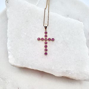 Peut inclure: Un collier doré avec un pendentif en forme de croix. La croix est ornée de plusieurs pierres précieuses roses rondes. Le collier est présenté sur une surface blanche texturée.