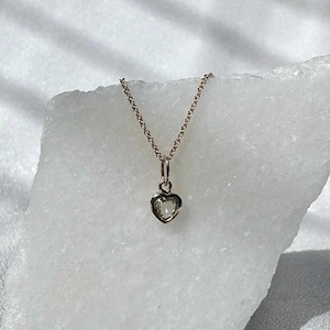 Puede incluir: Un delicado collar de cadena de oro con un pequeño colgante de corazón con una piedra preciosa transparente.