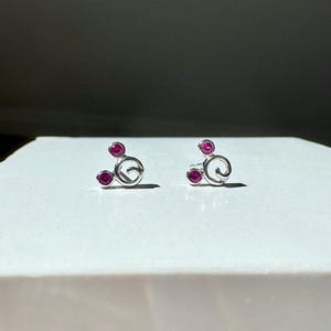 May include: A pair of silver-toned stud earrings. Each earring features a stylized design with three small, round, magenta-colored gemstones. The earrings are displayed on a white surface with soft lighting.