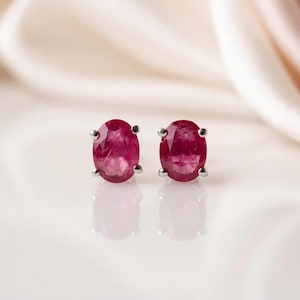 May include: A pair of oval-cut ruby stud earrings set in silver-toned metal prongs. Each earring features a deep red gemstone with visible internal facets and natural inclusions, reflecting clearly on the smooth surface below the jewellery pieces.