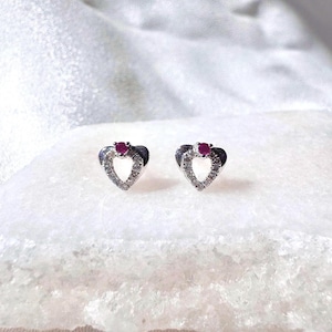 May include: A pair of heart-shaped stud earrings. Each earring features a heart outline encrusted with small, clear stones. A small, round, red gemstone is set at the top of each heart. The earrings are set against a white, textured background.