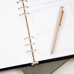 Op de afbeelding: Een witte planner met een zwarte kaft en gouden ringen. De planner heeft secties voor afspraken, vragen en plannen. Een gouden pen ligt op de open planner.