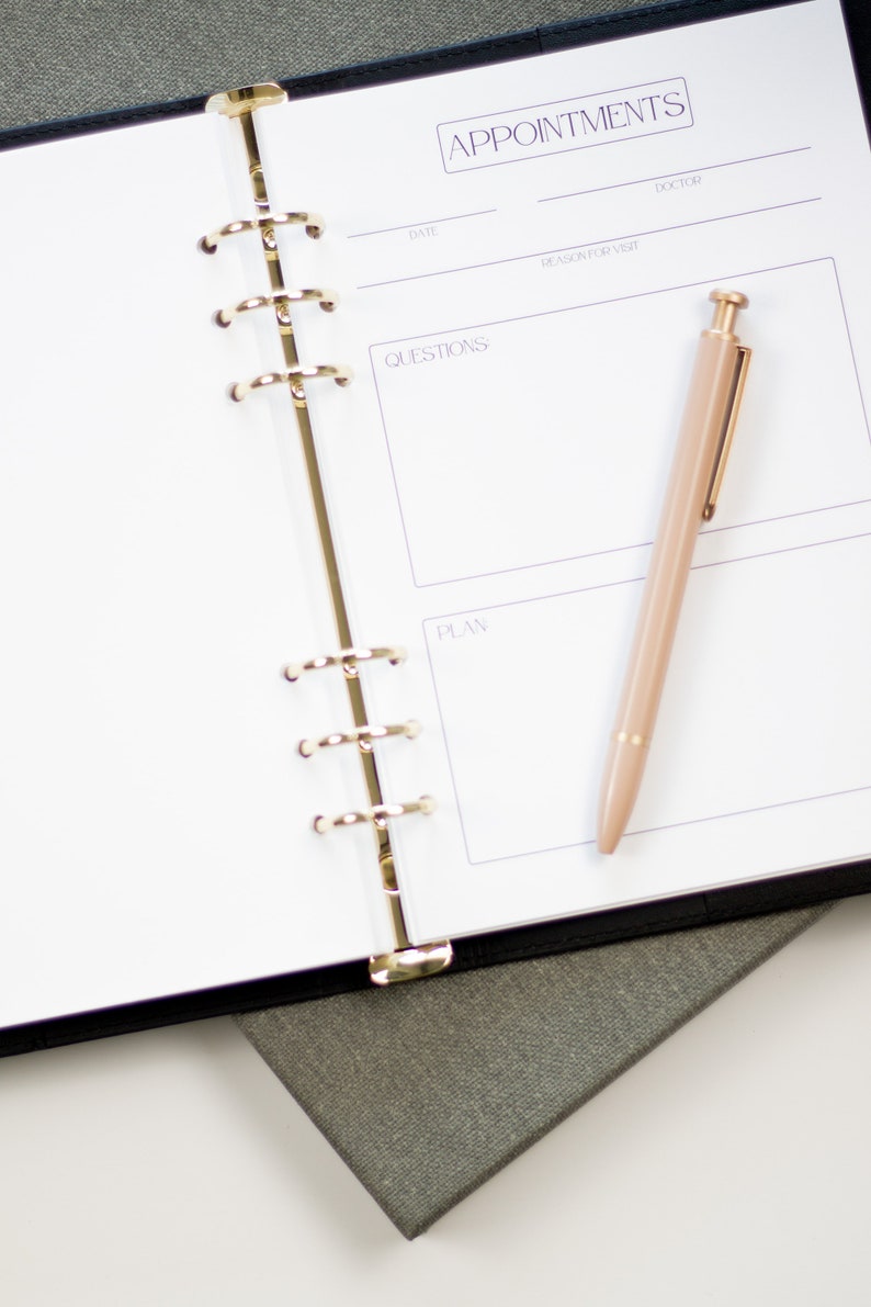 Op de afbeelding: Een witte planner met een zwarte kaft en gouden ringen. De planner heeft secties voor afspraken, vragen en plannen. Een gouden pen ligt op de open planner.