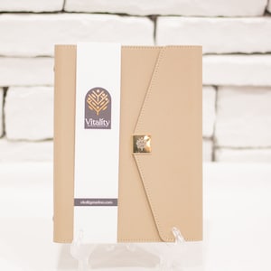 Op de afbeelding: Een beige leren planner met een gouden sluiting en een wit label met de tekst "Vitality Medical Record" en de website "vitalitymedrec.com".