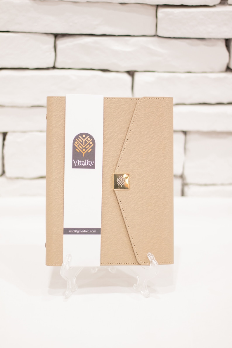Op de afbeelding: Een beige leren planner met een gouden sluiting en een wit label met de tekst "Vitality Medical Record" en de website "vitalitymedrec.com".