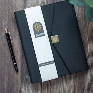 Puede incluir: Carpeta negra con aspecto de cuero con una banda blanca con el logotipo y el sitio web "Vitality Medical Record". Un emblema cuadrado dorado está en la cubierta. Un bolígrafo negro con detalles dorados descansa sobre una superficie de madera.