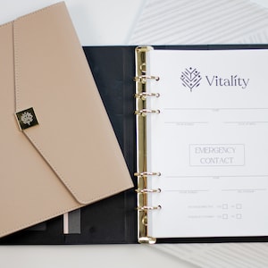 Op de afbeelding: Een zwarte en beige leren planner met gouden ringen en een witte pagina die open is naar een sectie voor noodcontacten. De planner heeft een logo met de tekst "Vitality" en een gestileerd boomontwerp.
