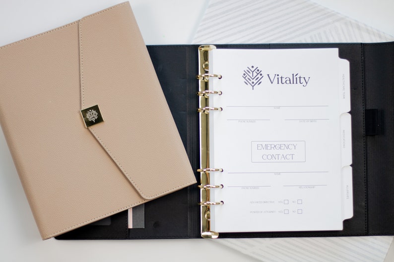Op de afbeelding: Een zwarte en beige leren planner met gouden ringen en een witte pagina die open is naar een sectie voor noodcontacten. De planner heeft een logo met de tekst "Vitality" en een gestileerd boomontwerp.