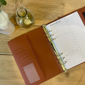 May include: Open brown leather daily planner with gold rings, a pen, and a "Daily Plan" page. The planner is open on a wooden surface, with a small glass bottle and a white flower arrangement in the background.