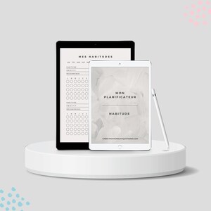 May include: Two digital tablets on a white pedestal. The tablet on the left has a black screen with the text "Mes Habitudes" and a habit tracker. The tablet on the right has a white screen with the text "Mon Planificateur Habitude" and the website "creerparmonsunquotidien.com".