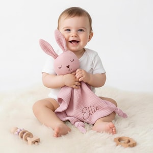 Könnte beinhalten: Eine rosa Baby-Schmusedecke mit einem Hasenkopf und dem Namen "Ellie" darauf gestickt. Der Hase hat geschlossene Augen und lange Ohren. Das Baby trägt einen weißen Body und sitzt auf einer weißen, flauschigen Oberfläche.