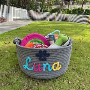 May include: A gray rope basket filled with dog toys, including a pink frisbee, a green bone-shaped toy, and a small can. The basket is personalized with the name "Luna" in colorful letters and a paw print.