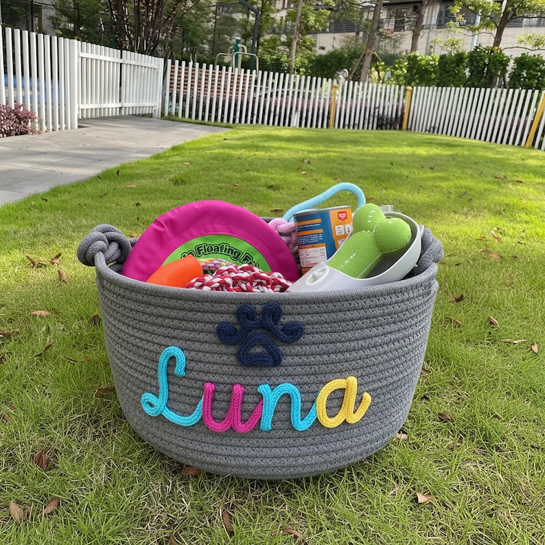 May include: A gray rope basket filled with dog toys, including a pink frisbee, a green bone-shaped toy, and a small can. The basket is personalized with the name "Luna" in colorful letters and a paw print.