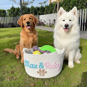 May include: Two dogs sit on grass next to a white rope basket filled with toys. The basket has the names "Max & Ruby" embroidered in blue and pink, with a paw print. One dog is golden, the other is white.