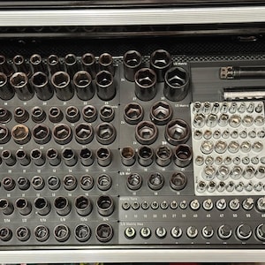 Könnte beinhalten: Eine Sammlung verschiedener Steckschlüssel und Werkzeuge, die ordentlich in einer Werkzeugkasten-Schublade angeordnet sind. Die Steckschlüssel sind in Reihen und Spalten angeordnet, mit Größen und Typen deutlich gekennzeichnet. Die Werkzeuge sind hauptsächlich dunkelgrau und silberfarben.