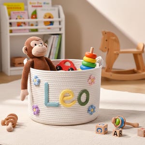 May include: A white rope basket with the name "LEO" in colorful letters, filled with toys including a stuffed monkey, a red car, and a wooden stacking ring. A wooden rocking horse and other toys are in the background.