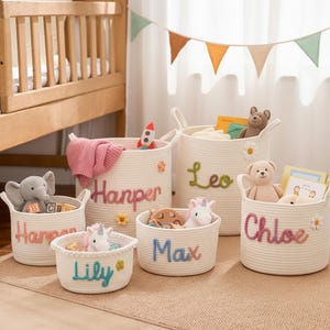 May include: A set of white rope storage baskets, each with an embroidered name and floral details. The baskets hold toys like a stuffed elephant, teddy bear, and unicorn. The names are in different colours, and the baskets sit on a neutral rug.