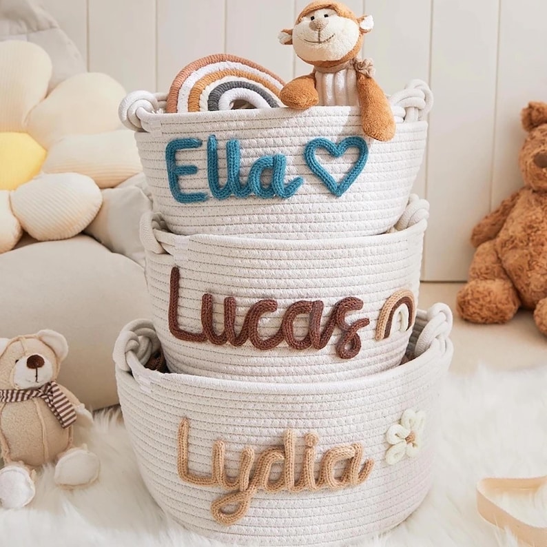 May include: Three white rope baskets stacked on top of each other. The top basket has the name "Ella" in blue letters and a blue heart. The middle basket has "Lucas" in brown letters. The bottom basket has "Lydia" in tan letters and a white flower.