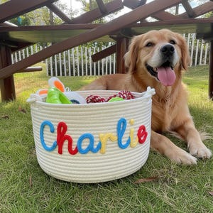 May include: A cream-colored rope basket with the name "Charlie" embroidered in blue, red, and yellow. The basket is filled with dog toys and sits on the grass next to a golden retriever.