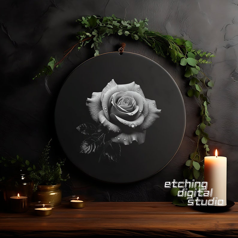 May include: A black and white illustration of a rose with water droplets on the petals. The illustration is on a round black canvas with a wooden frame. The canvas is hanging on a dark gray wall with greenery around it. There are candles and small jars on a wooden table in the foreground. The text "etching digital studio" is visible in the lower right corner of the image.