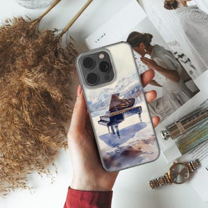 Può includere: Una custodia per telefono trasparente con un design ad acquerello di un pianoforte a coda in un paesaggio di montagna innevato. La custodia ha un rigonfiamento della fotocamera nero con tre obiettivi.