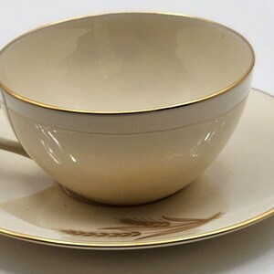 May include: A cream-colored teacup and saucer with a gold rim and a wheat design. The cup has a rounded shape and a handle.