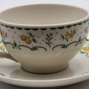 May include: Cream-colored teacup and saucer set. The cup and saucer are decorated with yellow floral and fruit designs, green scalloped trim, and a matching handle. The set is made of ceramic.