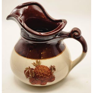 May include: A ceramic pitcher with a brown rim and handle, and a cream-colored body. The pitcher features a pineapple and leaf design in brown and tan. The handle has a decorative swirl.