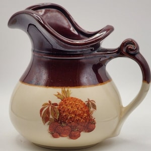 May include: A brown and cream ceramic pitcher with a pineapple and strawberries design. The pitcher has a wide, flared lip and a handle with a scroll design.