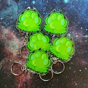 Op de afbeelding: Vijf heldere acryl sleutelhangers met een groen pootafdrukontwerp en zwarte accenten. Elke sleutelhanger heeft een zilverkleurige ketting en een ronde sleutelring. De achtergrond heeft een galaxy-ontwerp.