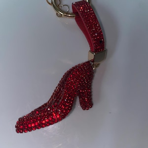 May include: A high-heeled shoe keychain covered in small, sparkling red crystals. The shoe is attached to a gold-toned metal ring by a matching red crystal-encrusted strap with a gold-toned metal connector piece at the top of the shoe.