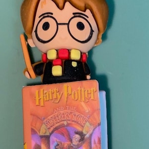 May include: A Harry Potter-themed bookmark featuring a cartoon figure with glasses, a wand, and a scarf. The bookmark is placed on a miniature book titled "Harry Potter and the Sorcerer's Stone."