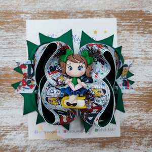 May include: Green and white hair bow featuring a girl doll sitting on a book. The doll is wearing a green shirt, blue skirt, and holding a book. The bow has a school theme with letters and stars.