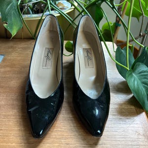 May include: A pair of black patent leather pumps with pointed toes. The shoes have a low heel and a V-shaped vamp. The brand name "Amalfi" is printed on the insole. The shoes are displayed on a wooden surface.