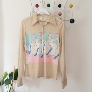 May include: A beige long-sleeved button-up shirt with a colorful print of women in hats and dresses. The print is on a light blue, pink, and white background.
