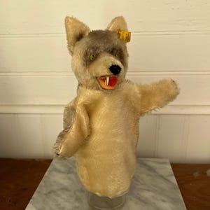 May include: A plush wolf hand puppet featuring beige and grey fur, green eyes, a black nose, and an open mouth with a red tongue and white teeth. A small yellow tag is attached to the ear with the text Made in Germany visible on the fabric.