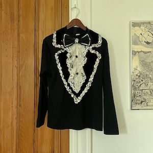 May include: Black long-sleeve top with a tuxedo-style design. The top features a black velvet bow tie and collar, with a white lace insert. Silver sequins outline the collar and the front of the top.