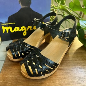 May include: Black leather sandals with a woven design on the toe and ankle straps. The sandals have wooden soles and heels. The background includes a record album with the text "Jacques Meuris" and the word "magri".