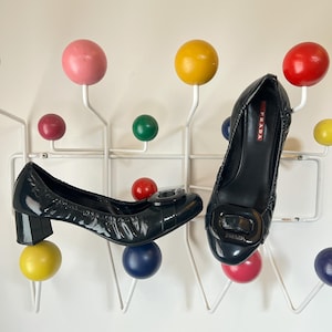 May include: Two black patent leather Prada pumps with square buckles. The shoes are displayed on a white coat rack with colorful wooden ball accents. The heels are approximately 2 inches high.