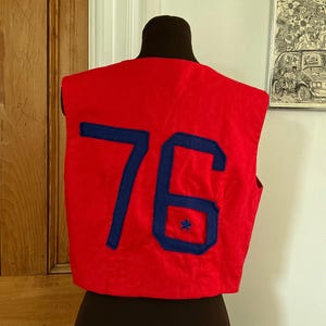 May include: A red vest with the number "76" in blue felt. A small blue star is embroidered inside the number "6". The vest is displayed on a mannequin.