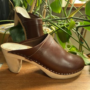 May include: A pair of brown leather clogs with wooden heels. The clogs have a rounded toe and a strap across the top. The heels are approximately 3 inches tall. The clogs are sitting on a wooden surface.