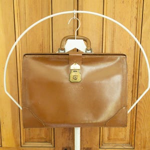 May include: A brown leather briefcase with a brass clasp and handle. The briefcase is hanging on a white hanger. The initials "G.W.P." are visible on the front of the briefcase.