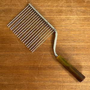 May include: A vintage cake slicer with a stainless steel wire grid and a brown and yellow handle. The slicer is designed to cut even slices of cake or other baked goods. The handle is made of a durable material.
