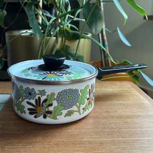 May include: White enamel saucepan with a black handle and lid. The pot features a floral design in shades of blue, green, and gold. The lid has a black knob. The saucepan is sitting on a wooden surface.