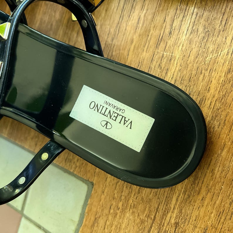 May include: A black Valentino Garavani sandal featuring a glossy finish, thin straps, and metallic studs. The footbed displays a white rectangular label with the brand name Valentino Garavani printed in black text, centered on the heel area.