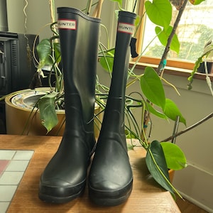 May include: A pair of black Hunter rain boots. Each boot has a red and white rectangular label near the top with the word "HUNTER" printed on it. The boots are tall, reaching up to the mid-calf. They are made of a matte rubber material.