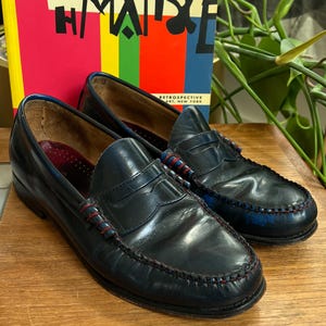 May include: A pair of blue leather penny loafers with red stitching and accents. The shoes are displayed on a wooden surface, with a colorful book in the background. The book has the word "MAISE" on the cover.