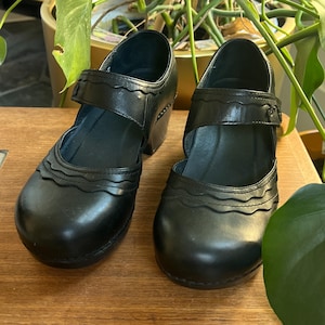 May include: A pair of black leather Mary Jane style shoes with a strap and buckle closure. The shoes have a rounded toe and a block heel. The upper features a decorative wavy design.