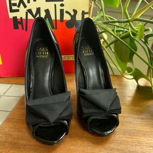 May include: A pair of black patent leather peep-toe heels with bow accents. The heels are labeled "SAKS FIFTH AVENUE" on the insole. The shoes are displayed on a wooden surface.
